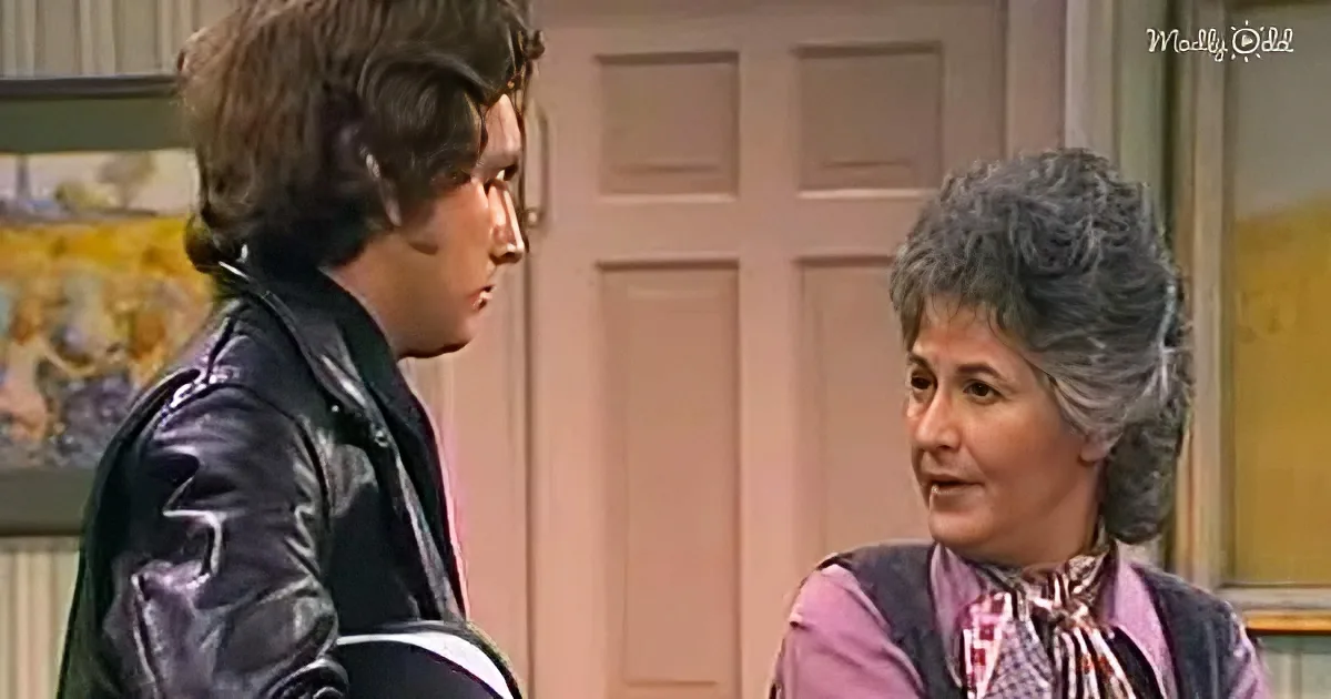 A man in a leather jacket and a woman in a floral blouse are having a conversation in a room with a door and artwork.