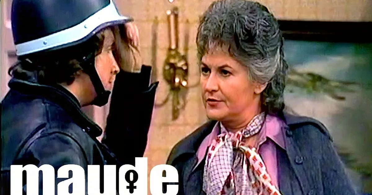 A woman in a purple jacket and checkered scarf stands beside a person in a black uniform, both facing each other.
