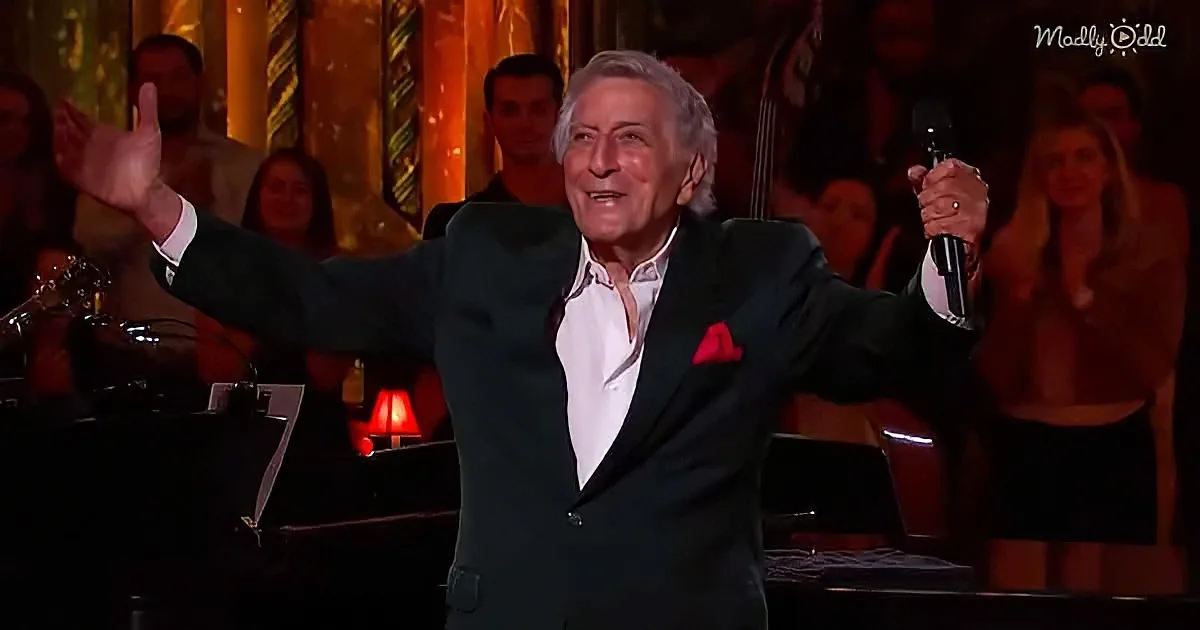 A man in a suit holds a microphone, gesturing with his right hand, in front of a live audience.