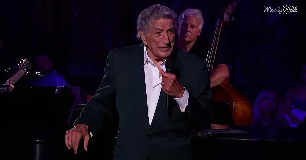 An older man in a suit holds a microphone, gesturing with his other hand on stage.