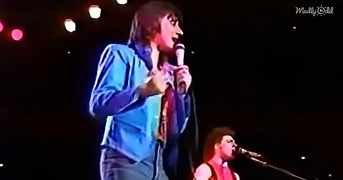 A person in a blue shirt and tie sings into a microphone on stage, with another performer in the background.