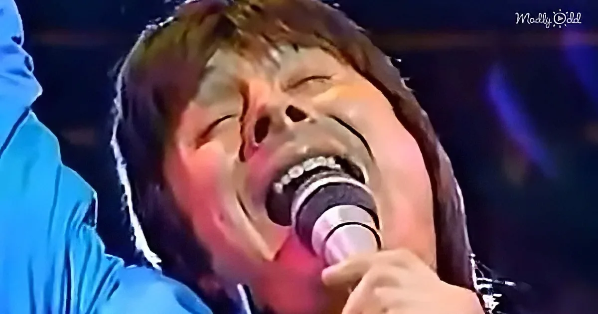 A person with long hair and a beard sings passionately into a microphone, illuminated by stage lighting.