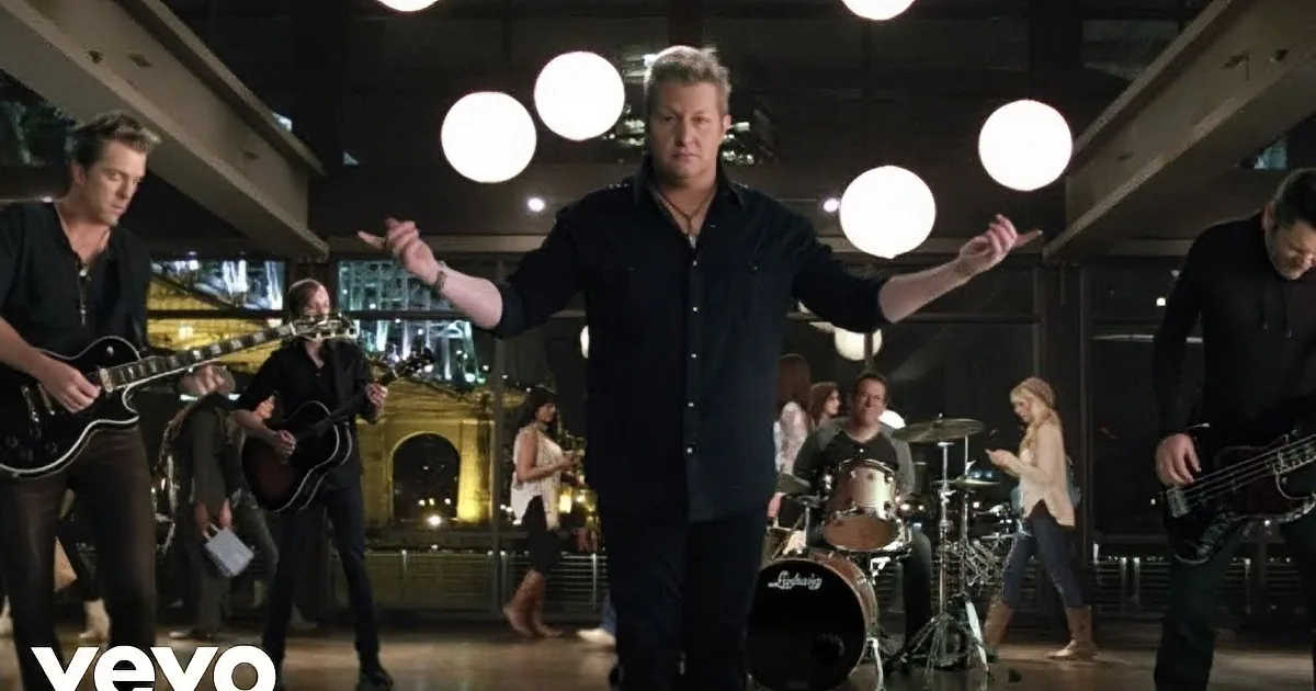 A man in black shirt and jeans stands with arms outstretched, singing in front of a band.