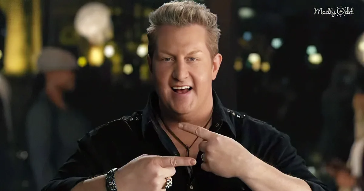 A man with blonde hair, wearing a black shirt and a necklace, points to his chest with both hands.