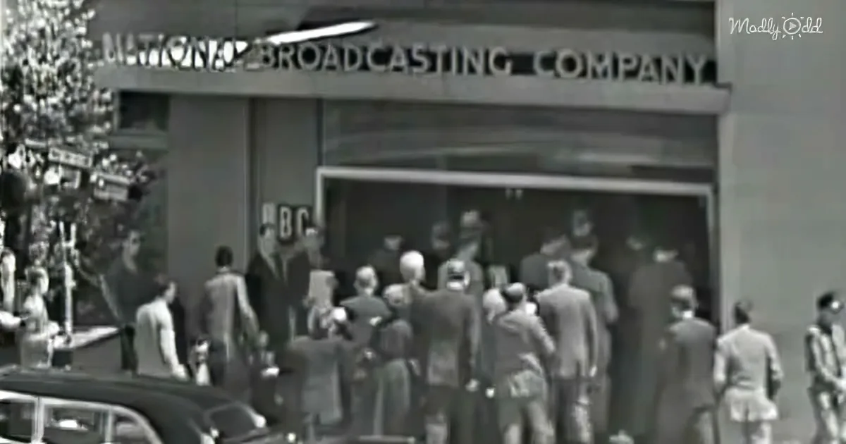 A crowd gathers outside the NBC building, with people in suits and hats, vintage cars nearby.