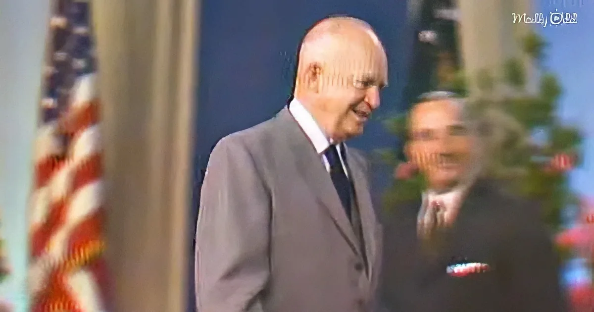 A man in a suit and tie smiles, with an American flag and another person in the background.