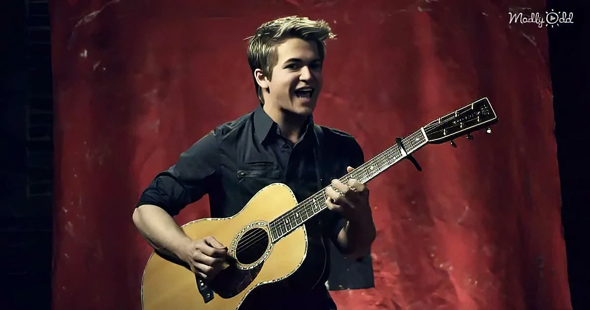 A man in a black shirt plays an acoustic guitar on stage with a red backdrop.