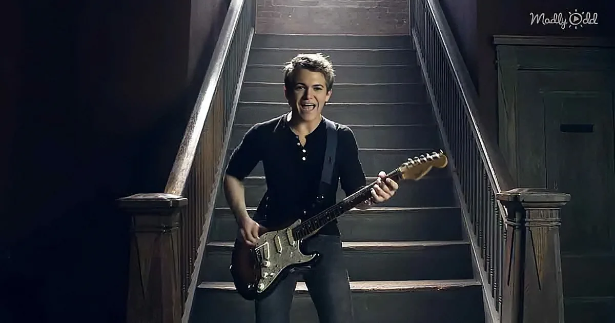 A person plays a guitar on a staircase, smiling, with a dark background and wooden railings.