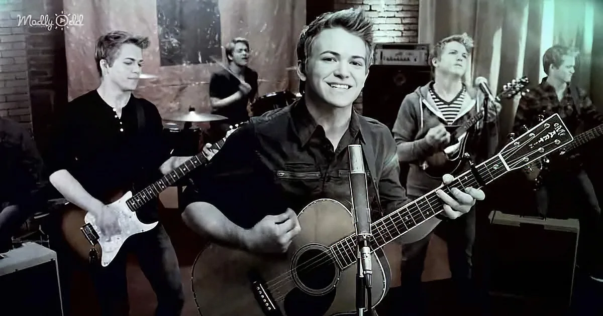 A smiling young man plays guitar in front of a band, with a microphone and a brick wall backdrop.
