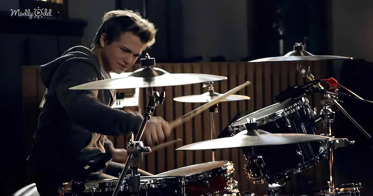 A young man in a hoodie is intensely playing a drum set in a dimly lit room.