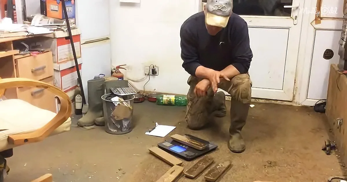 A man in a workshop squats, wearing a mask and gloves, surrounded by tools and materials on the floor.