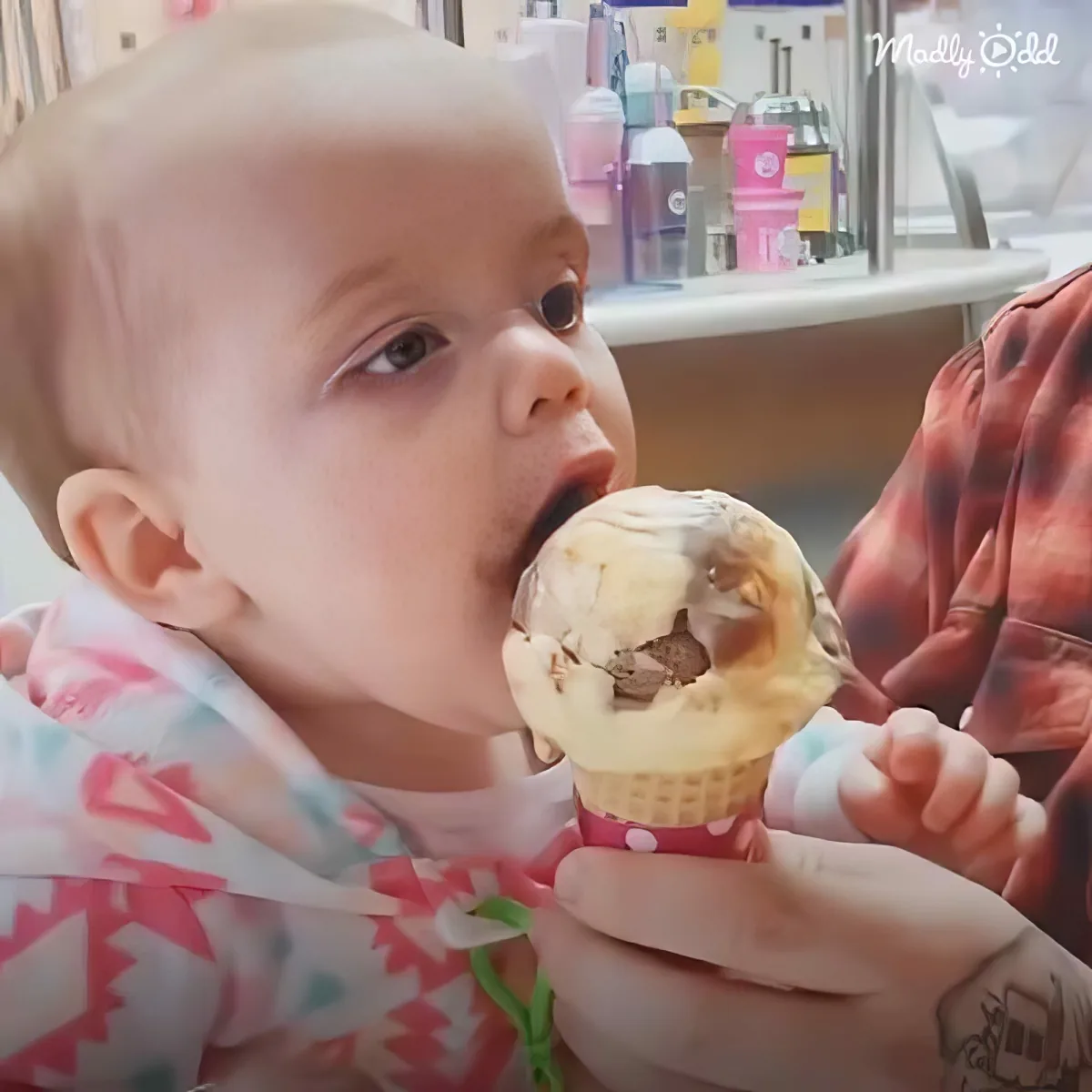 A baby in a colorful outfit enjoys a chocolate ice cream cone with a hand holding it.