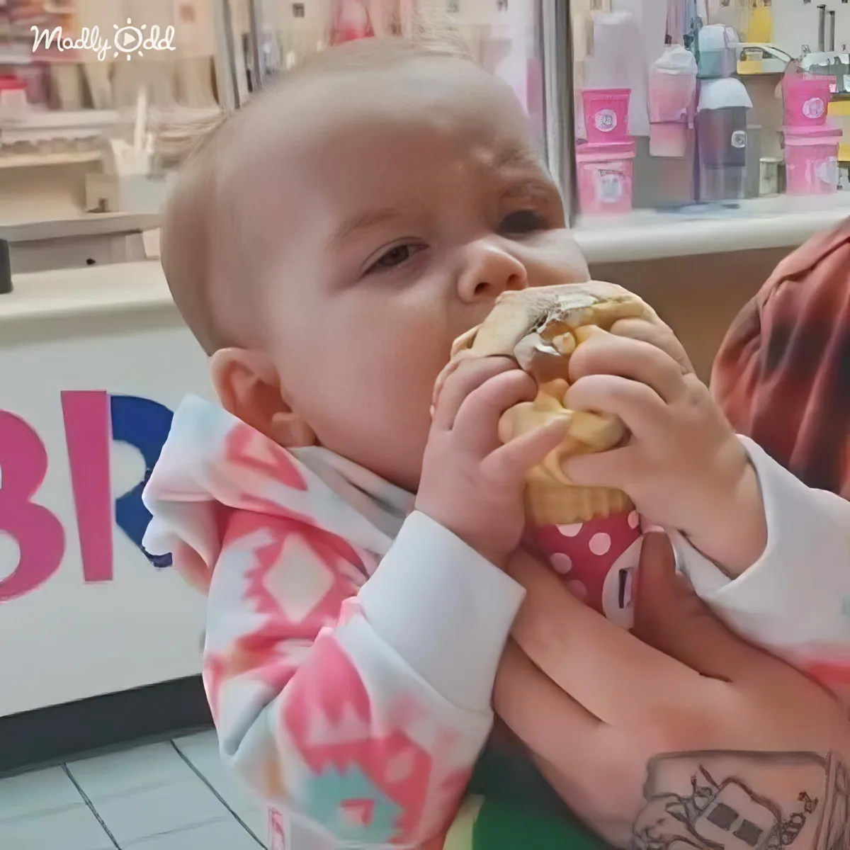 A baby in a colorful outfit is being held and eating an ice cream cone in a brightly lit setting.