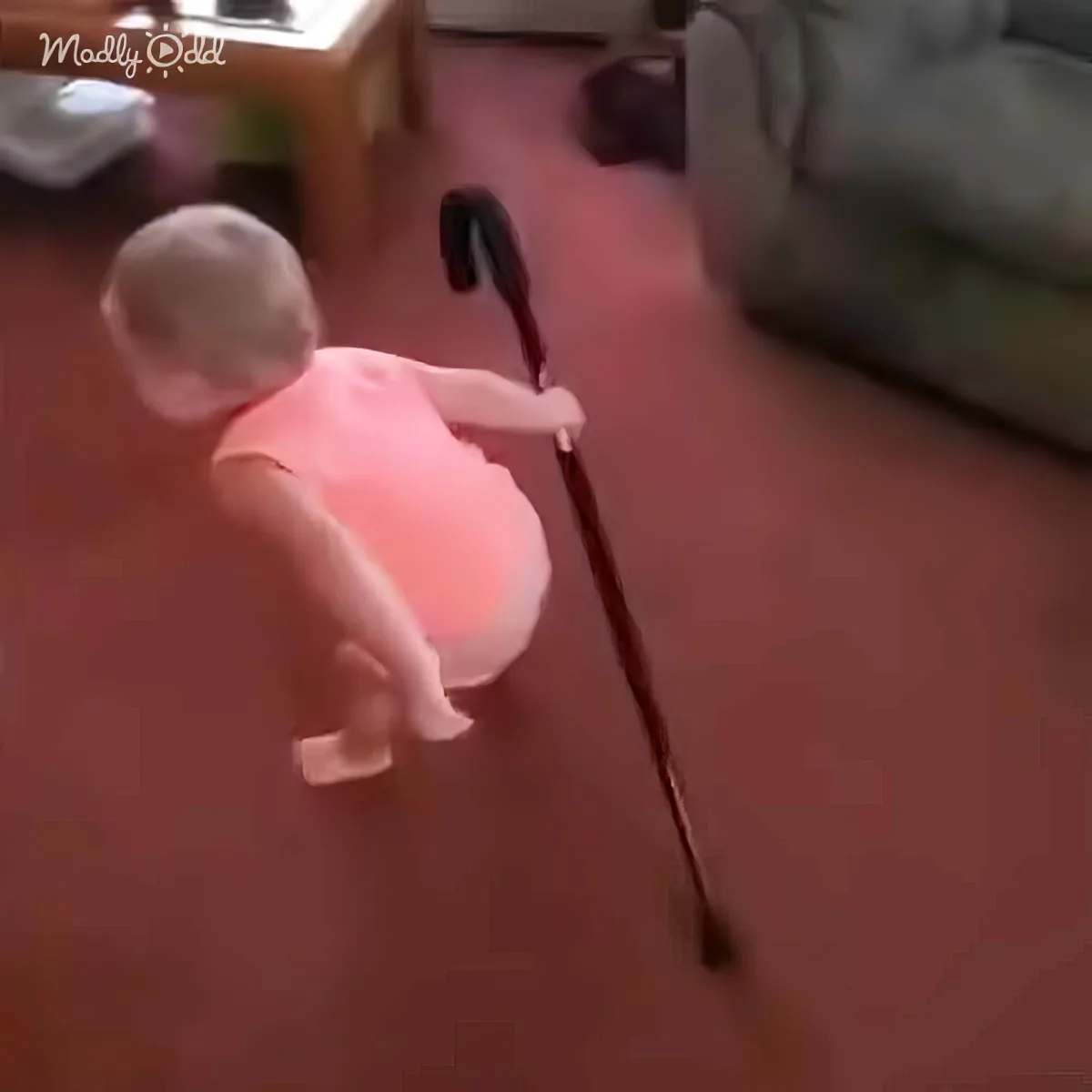 A baby in a pink outfit is holding a cane, standing on a red floor.