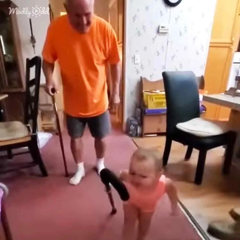 A man in an orange shirt uses a cane while a child holds a microphone, both in a home setting.