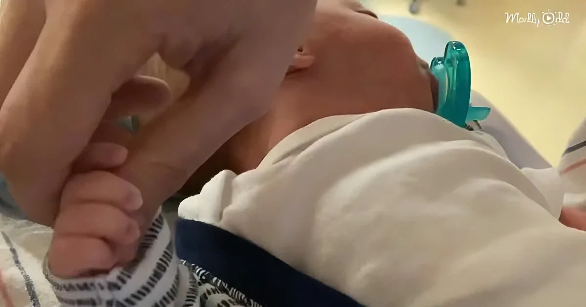 A newborn baby, wearing a white outfit with a blue stripe, is being bottle-fed while wearing a blue pacifier.