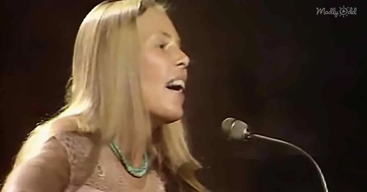 A woman with long blonde hair sings into a microphone, wearing a lace top and a necklace.