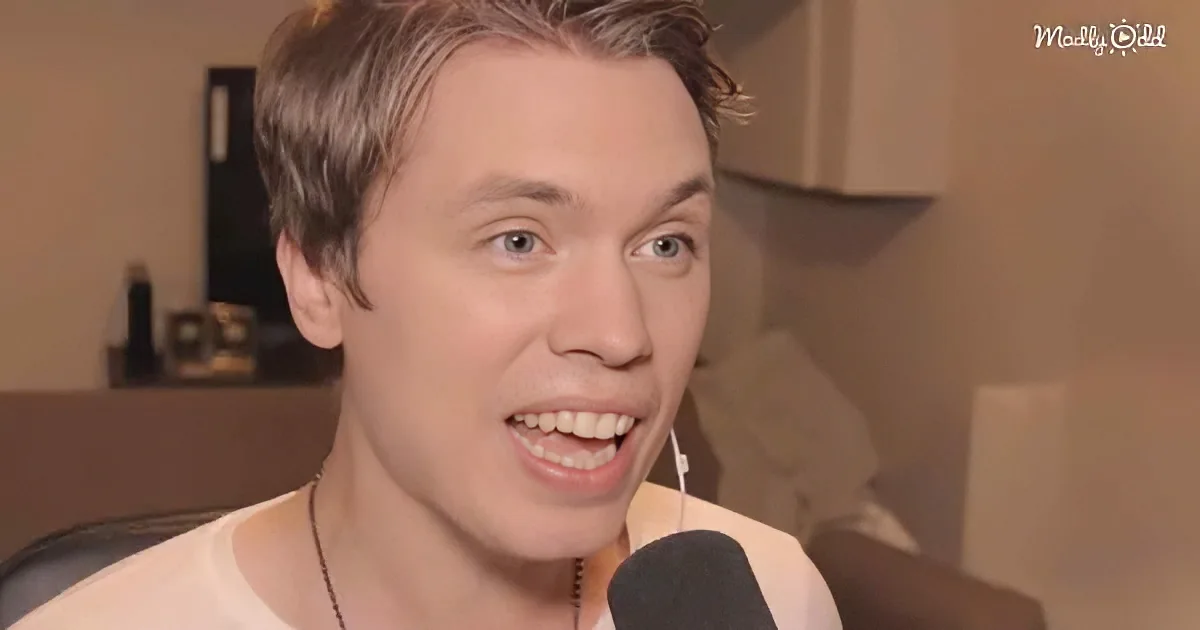 A person with short hair, wearing a white shirt, smiles while speaking into a microphone in a room with a blurred background.