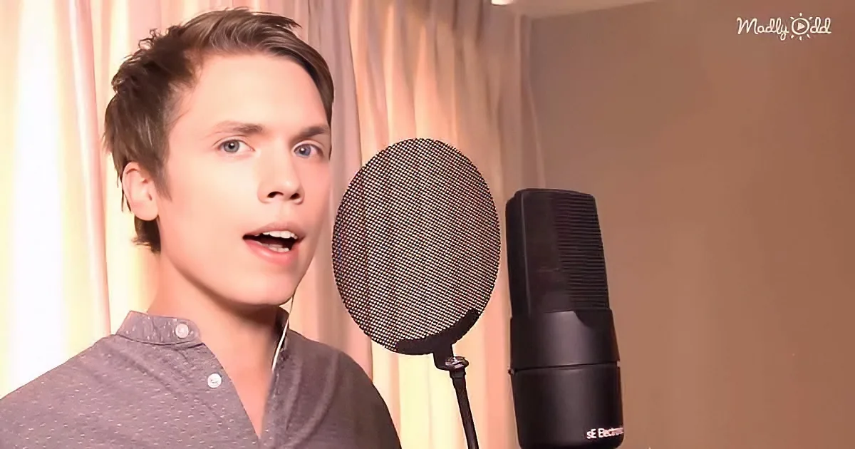 A person wearing a gray shirt sings into a microphone with a pop filter in a room with curtains.