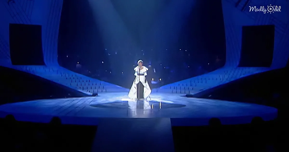 A person in a white outfit stands on a stage with blue lighting, surrounded by darkened audience areas.