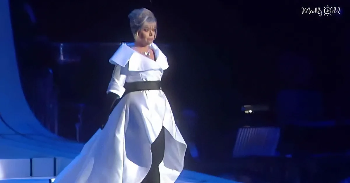 A woman in a white, flowing gown with a black belt stands on stage, illuminated by blue lighting.