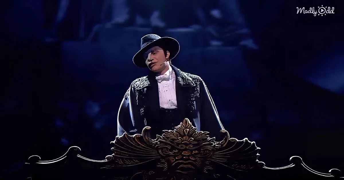 A person in a black suit and hat stands on stage, holding a ornate, dark-colored object.