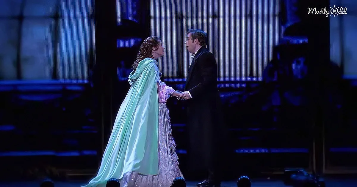 A woman in a green gown and a man in a suit hold hands on stage, illuminated by blue lighting.