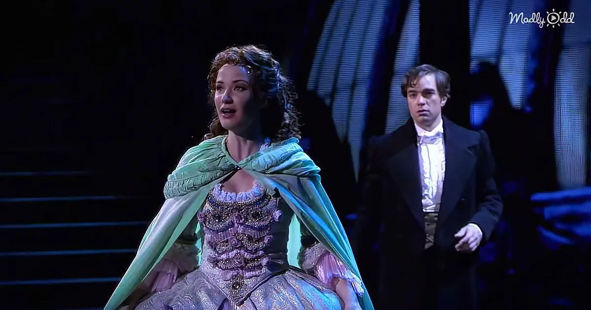 A woman in a purple dress with a green cape sings on stage, accompanied by a man in a tuxedo.