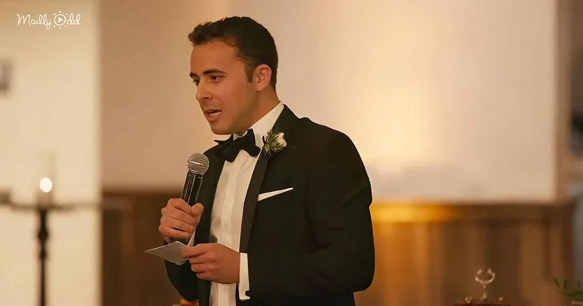 A man in a tuxedo holds a microphone and paper, speaking at a formal event.