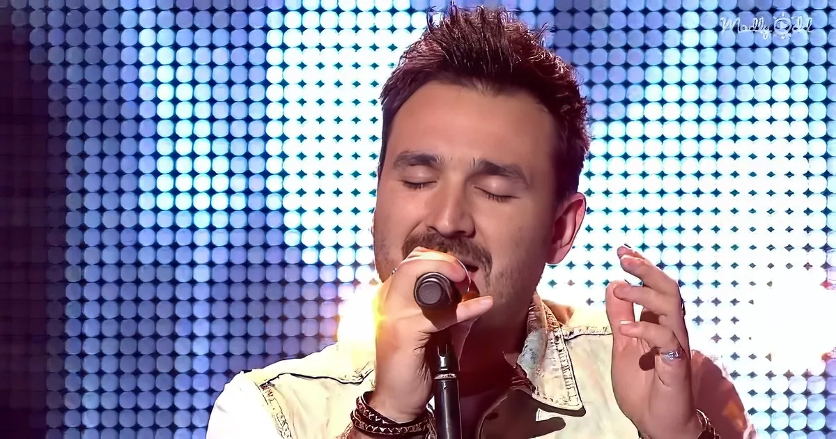 A man with closed eyes holds a microphone, wearing a denim jacket, in front of a bright, dotted background.