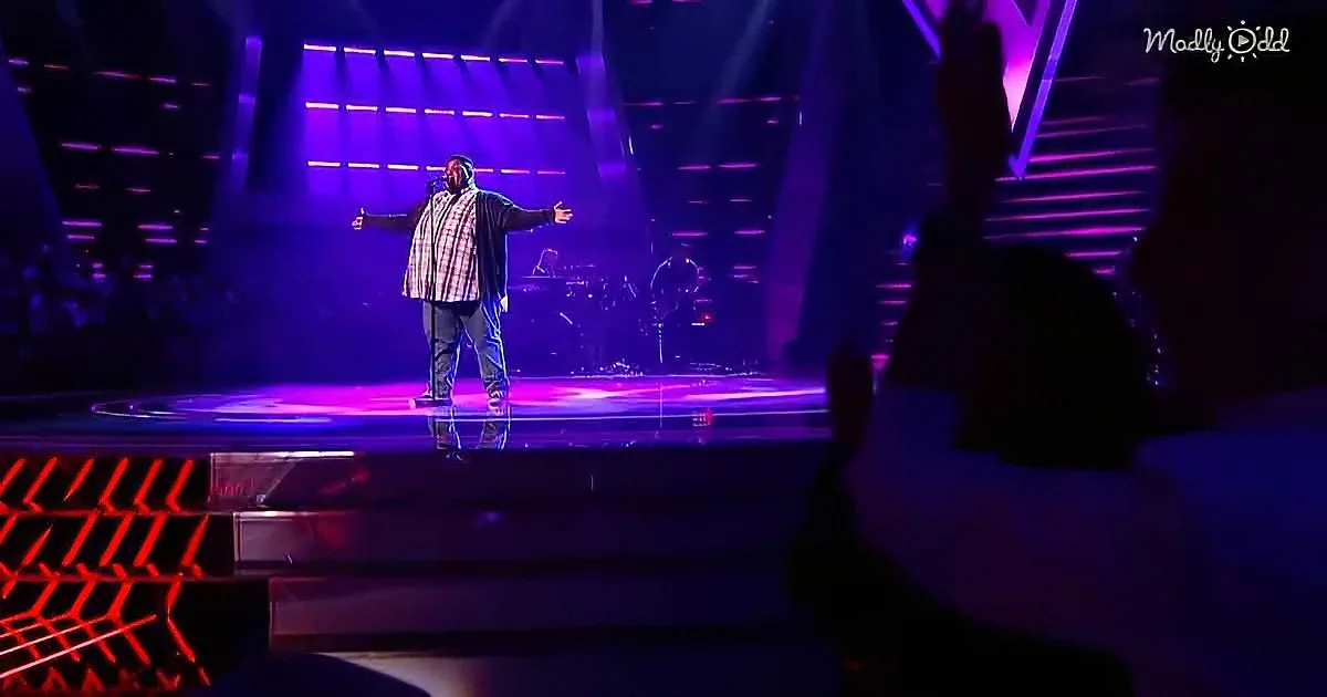A man stands on a stage with arms outstretched, bathed in purple light, with a band and audience in the background.