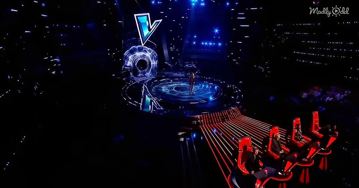A singer stands on a circular stage with a glowing blue "V" logo, surrounded by judges in red chairs.
