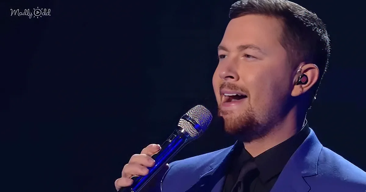 A man in a blue suit sings into a microphone, wearing an earpiece, against a dark background.