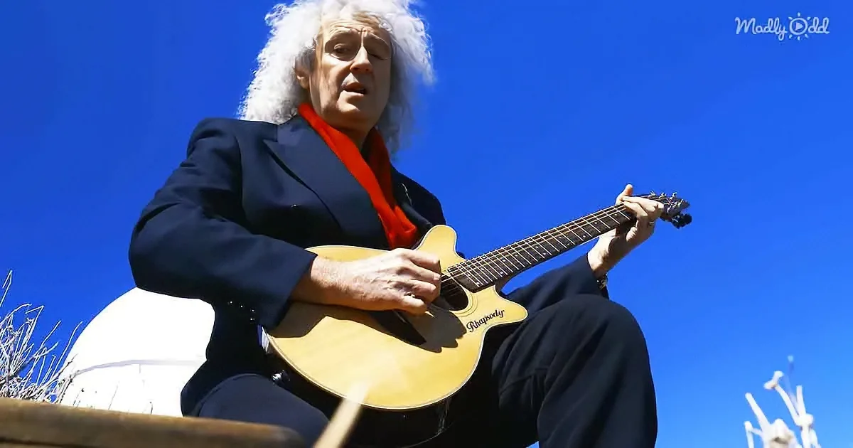 A man with curly hair in a suit plays a guitar against a clear blue sky.