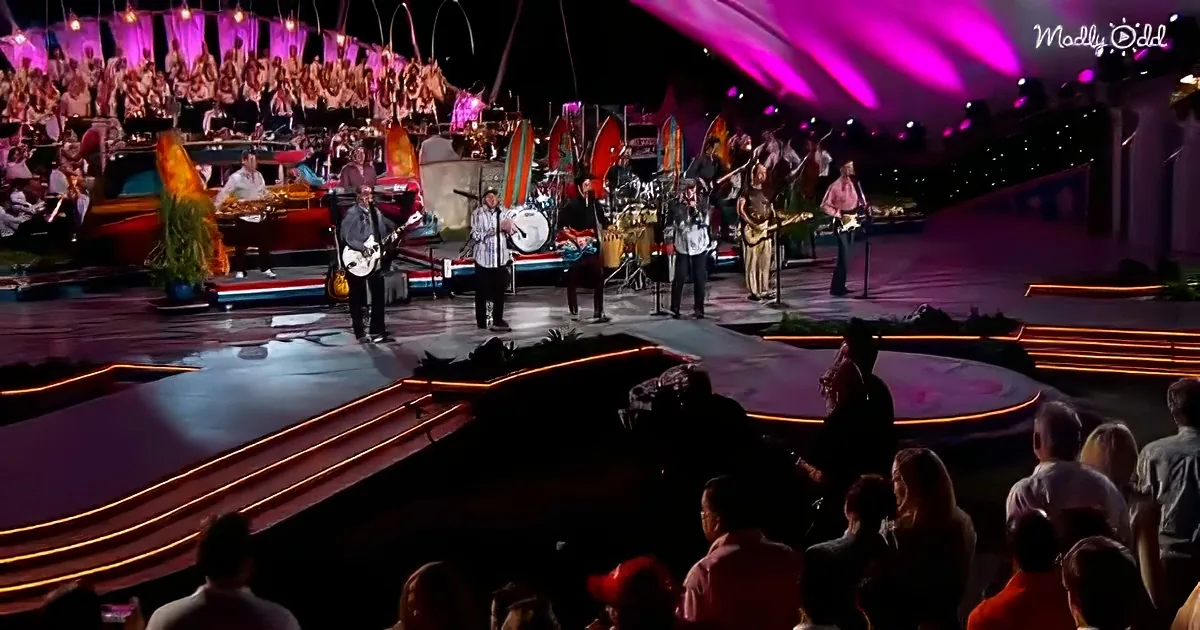 A band performs on stage with a large orchestra in the background, illuminated by vibrant purple lights.