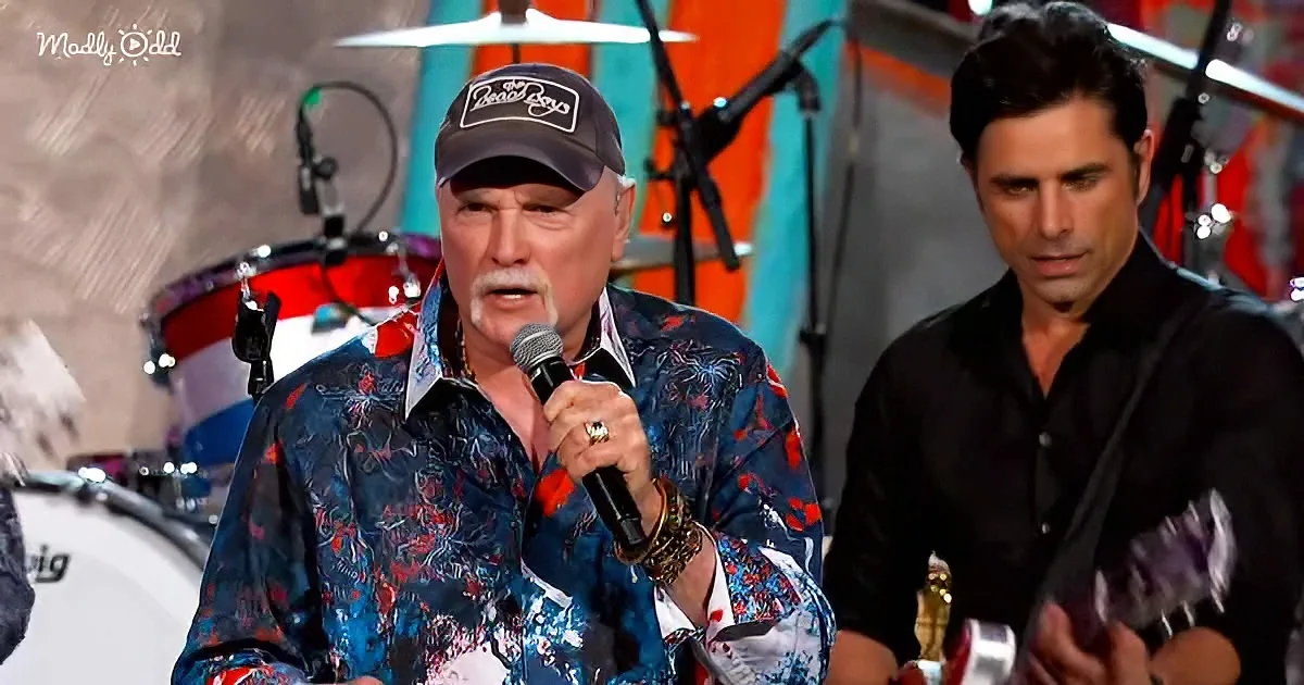 A man in a colorful shirt and cap sings into a microphone on stage, with another man in black standing nearby.