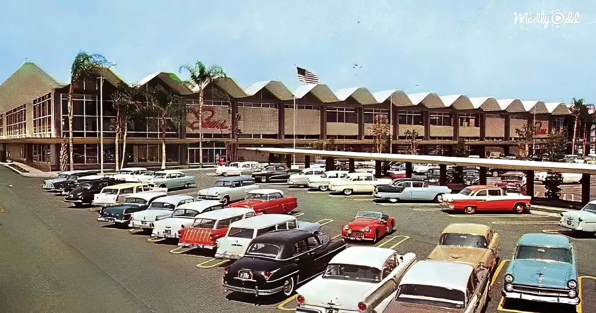 A bustling 1950s-era shopping mall with vintage cars parked in front, palm trees, and a clear blue sky.
