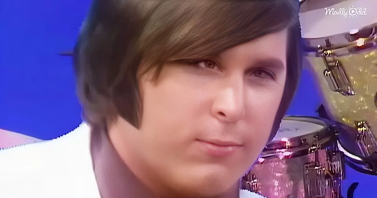 A person with short hair and a white shirt is in front of a drum set, with a blue background.