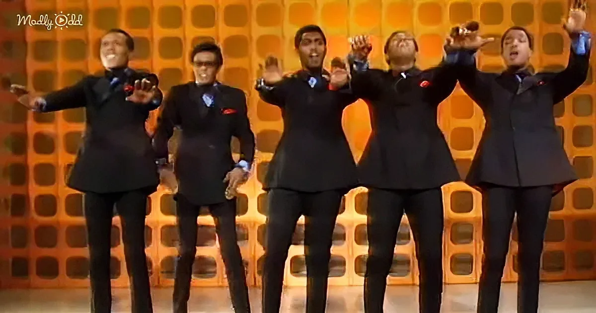 A group of five men in black suits with red accents pose with raised hands against a vibrant orange geometric background.