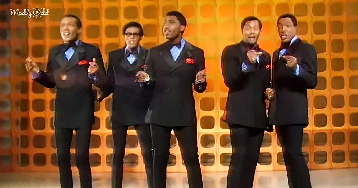 A group of five men in suits perform on stage, holding microphones and singing.