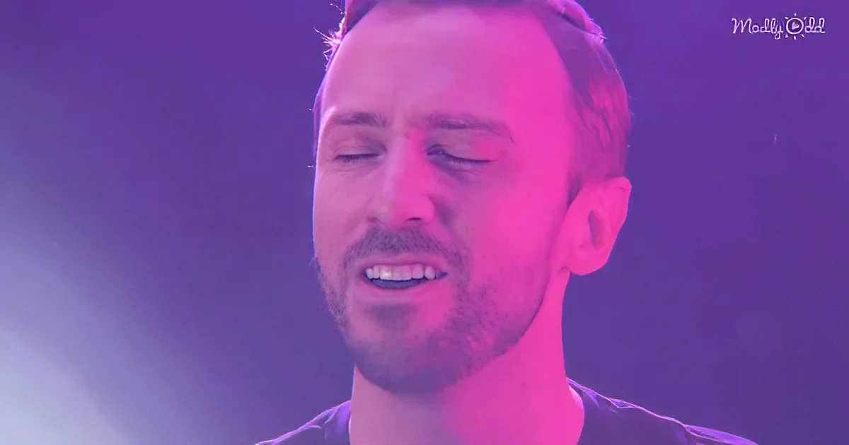 A man with closed eyes, wearing a black shirt, under a purple-lit background.