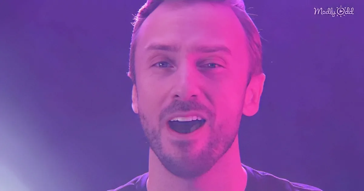 A man with short hair and a beard, wearing a dark shirt, is shown in a close-up with a purple and pink light effect.