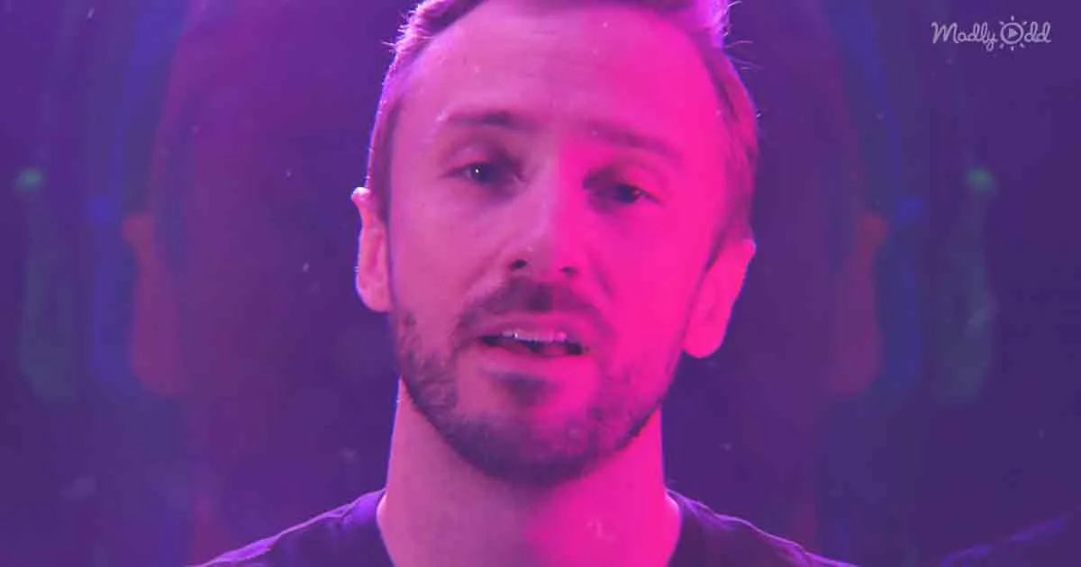 A man with short hair and a beard, wearing a dark shirt, stands under vibrant purple and pink stage lighting.