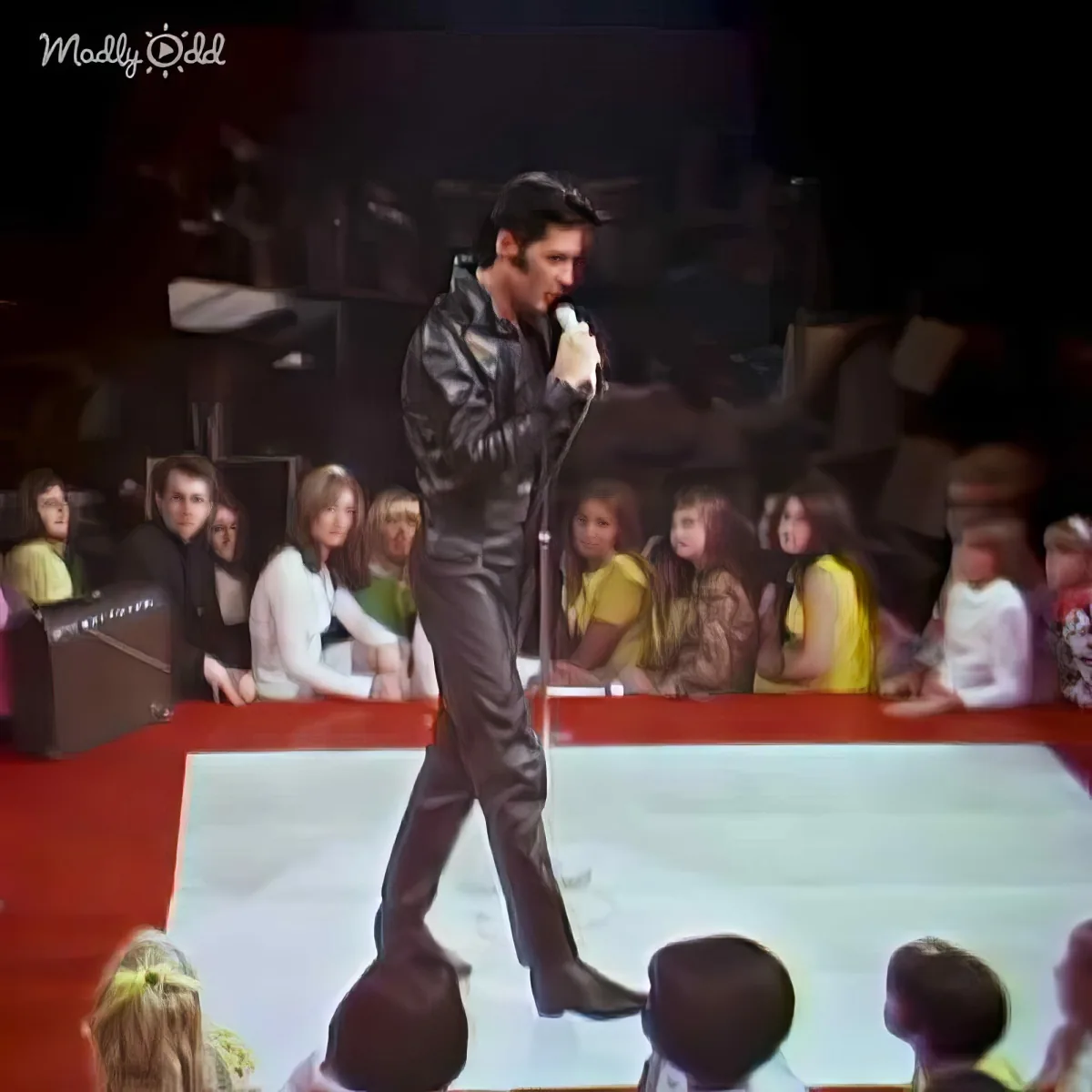 Elvis Presley in a black leather outfit sings on stage with a microphone, surrounded by an audience.