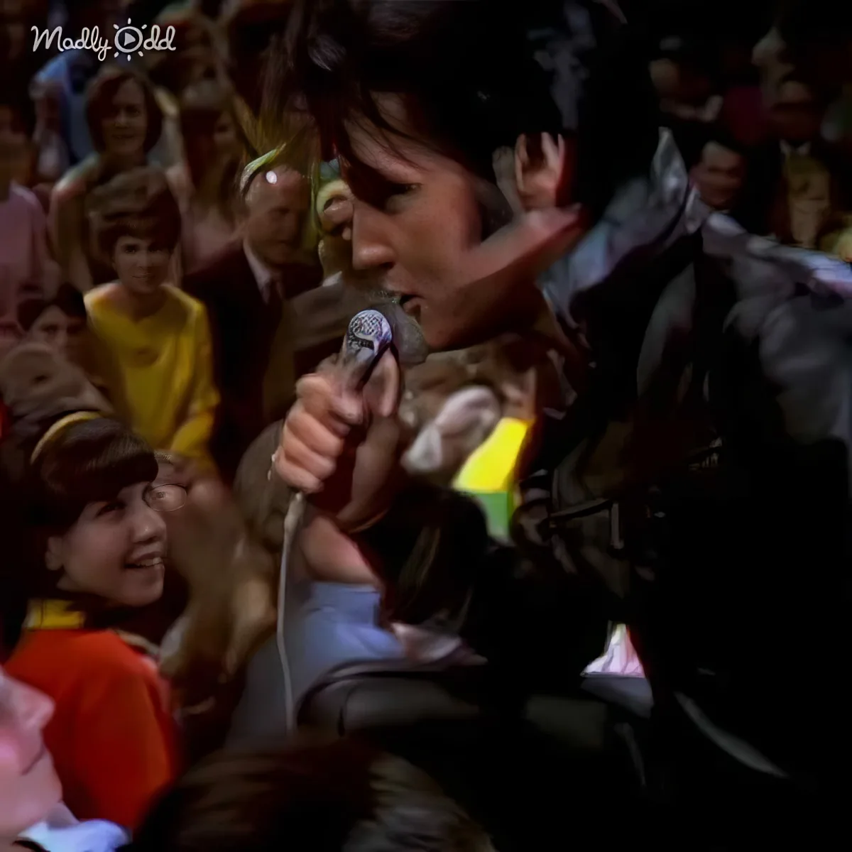 A man in a black outfit sings into a microphone, surrounded by a crowd of people in a lively setting.
