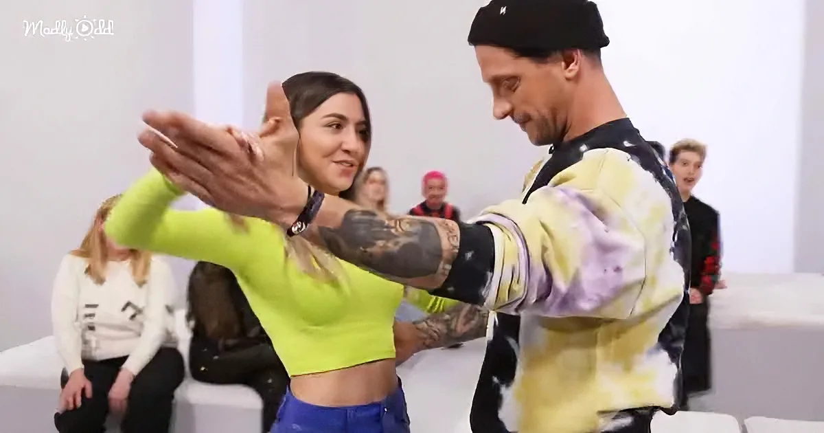 A man and woman are dancing, with the man's arm extended and the woman smiling, surrounded by onlookers.