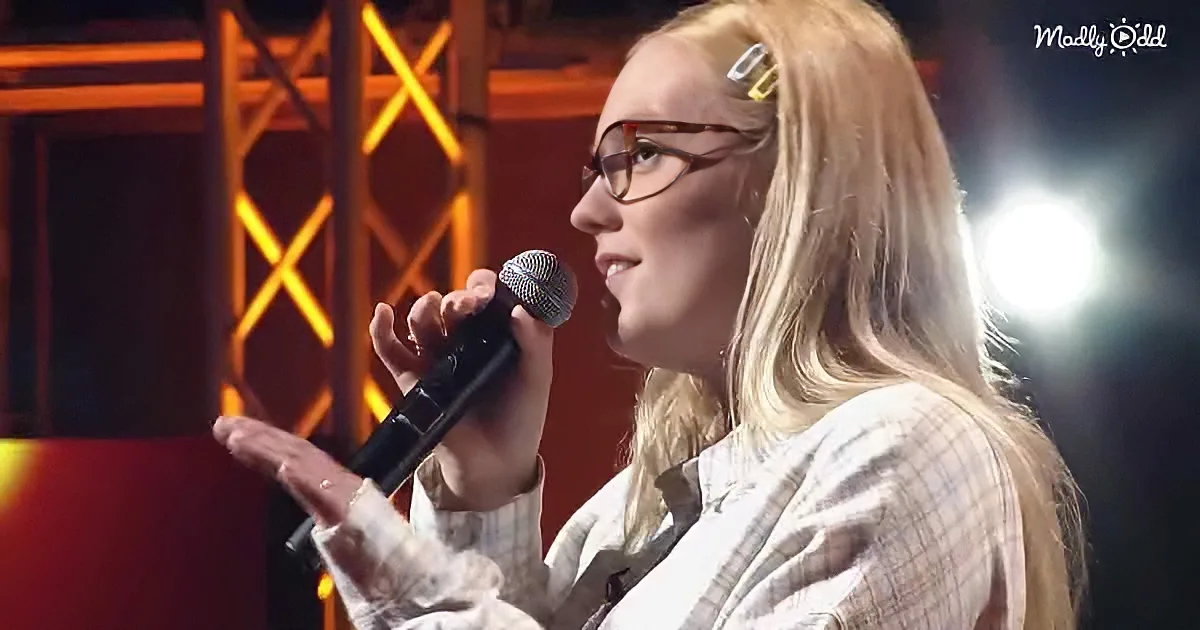 A person with long blonde hair and glasses holds a microphone, gesturing with their other hand.