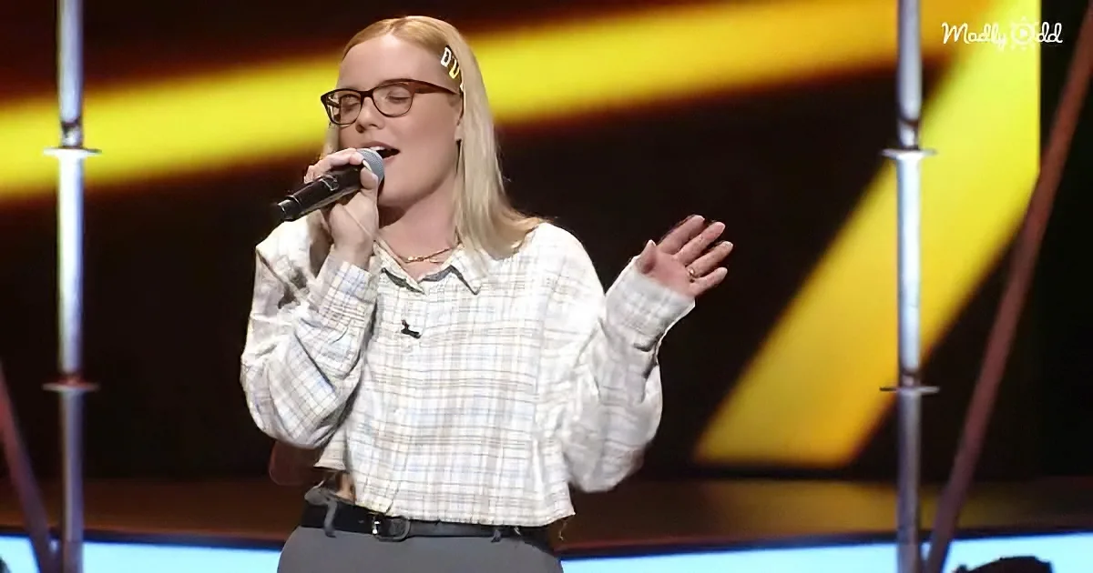 A person in a plaid shirt sings into a microphone on stage with a yellow and black background.