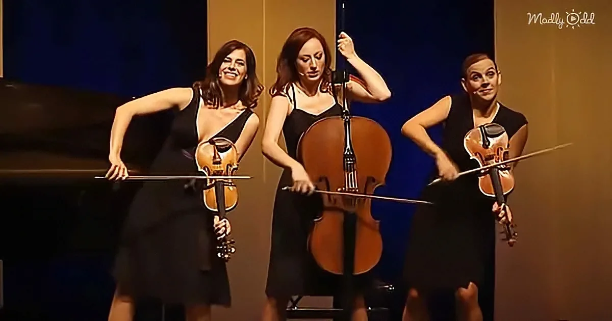 Three musicians in black dresses play string instruments on stage, smiling and holding their bows.
