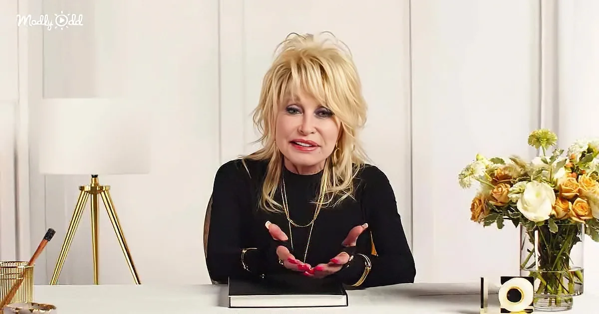 Dolly Parton sits at a table with a bouquet of flowers, wearing a black top and gold jewelry, gesturing with her hands.